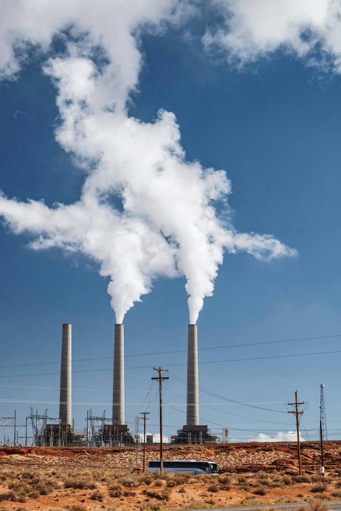 Tall smokestacks at an industrial plant with smoke against a blue sky.